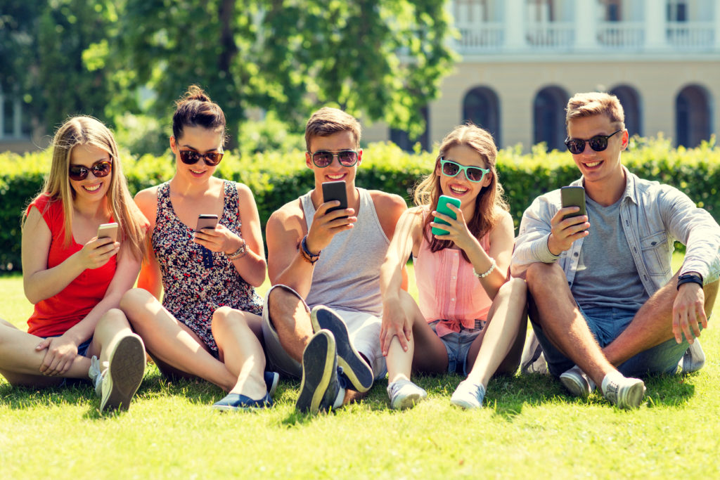 smiling friends with smartphones sitting on grass